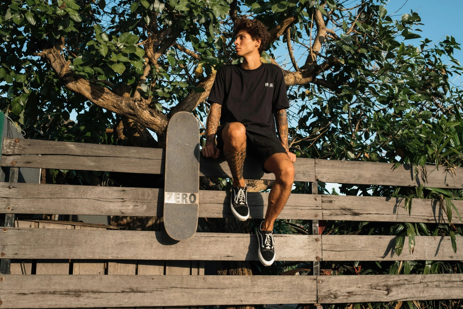 Teenager sitting on a wooden fence with a skateboard, enjoying the summer outdoors.