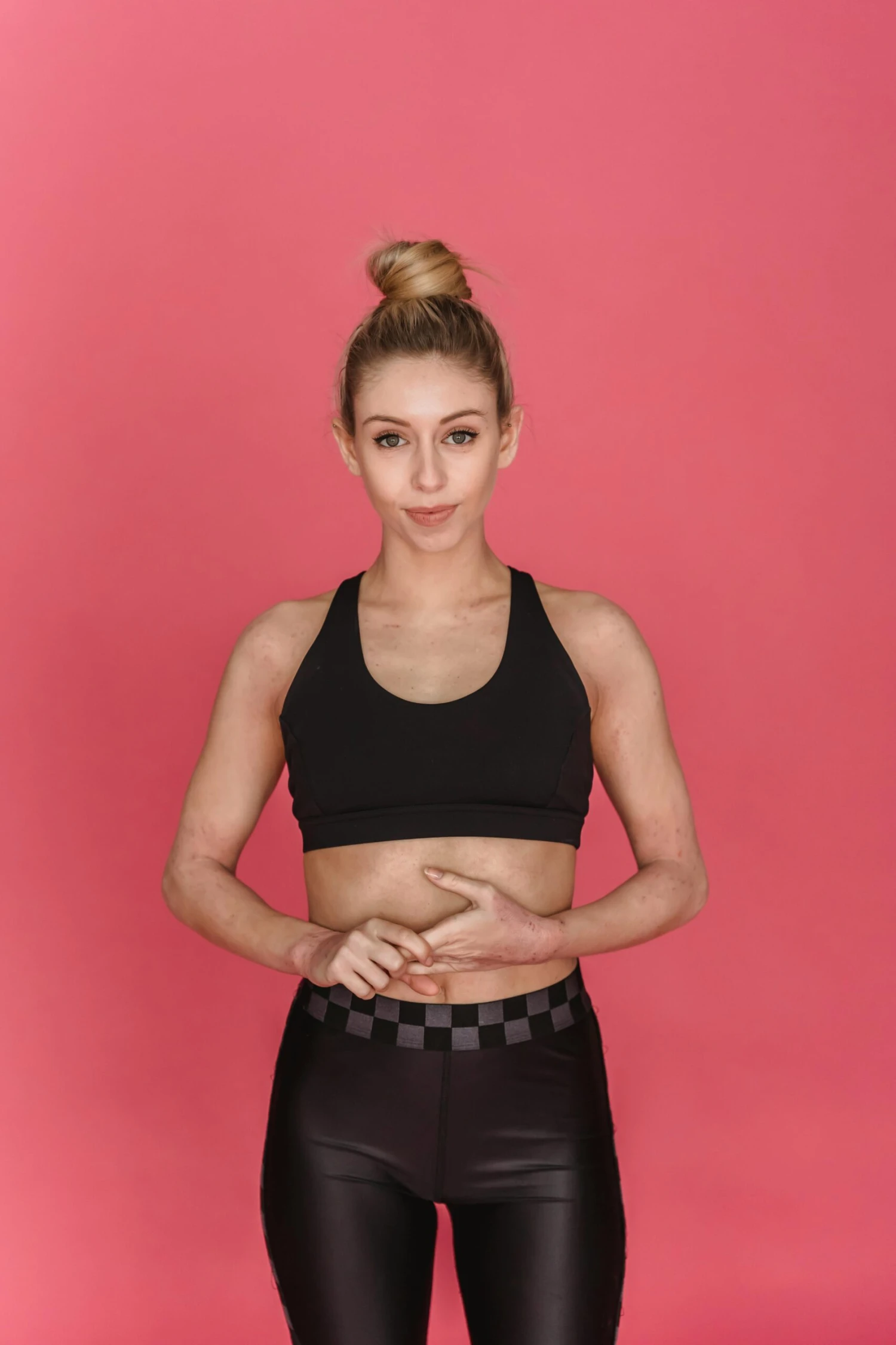 Young female athlete wearing black sports bra and leggings standing in studio against pink background and looking at camera